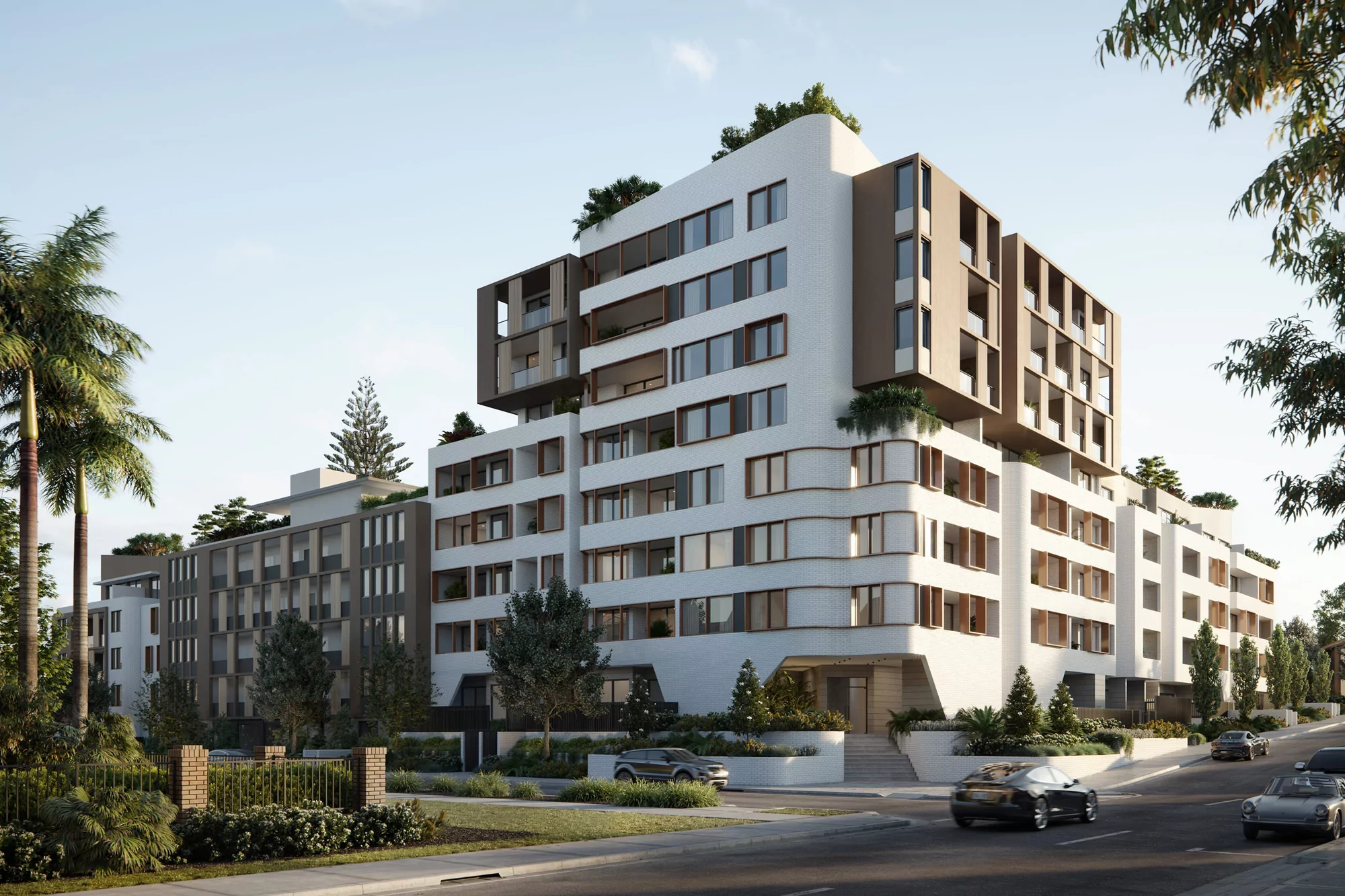Exterior of a modern residential building with white walls and brown accents, surrounded by trees and greenery, on a sunny day.