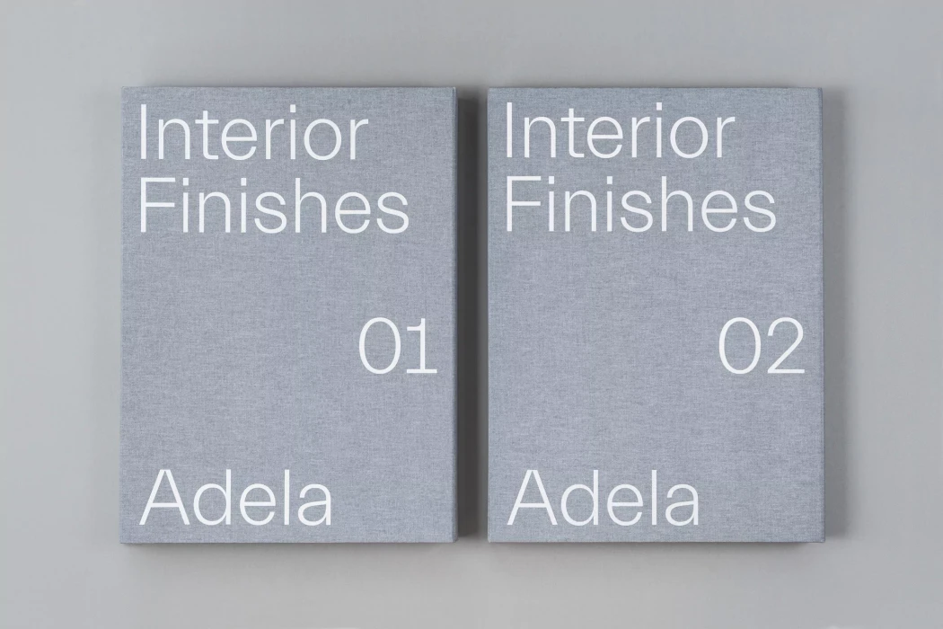 Two grey fabric-bound books with white typography titled Interior Finishes 01 and 02 for Adela on a neutral background