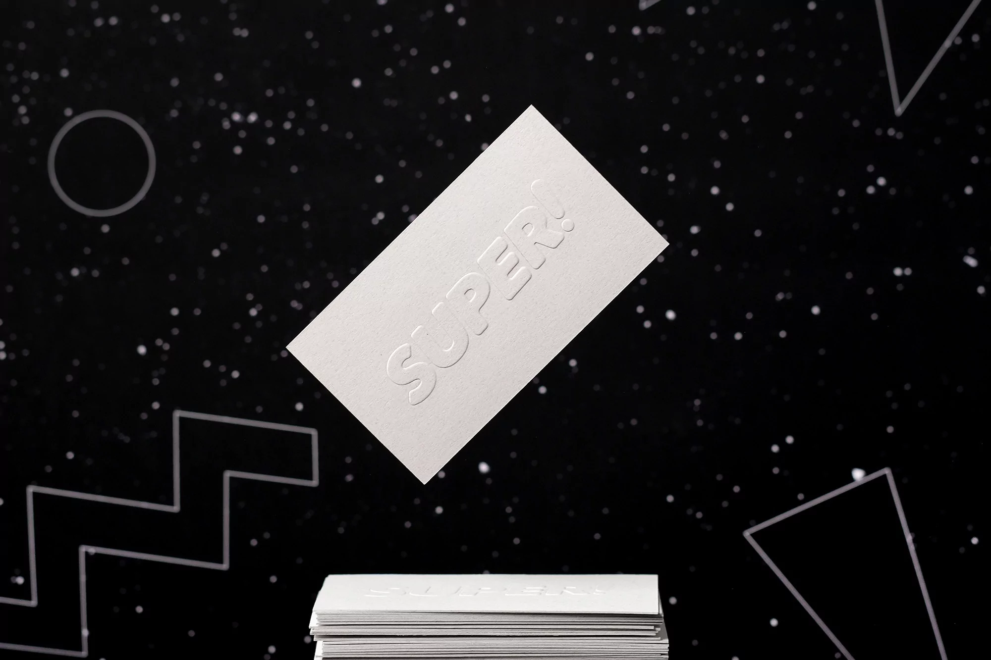 Studio shot of embossed business cards with the word 'SUPER!' stacked against a black background with white dots and line art.