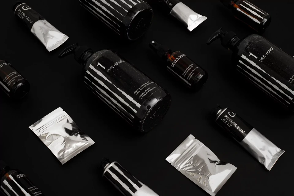 Several black and white cosmetic bottles and containers are arranged on a solid black background, some bottles have pumps and spray nozzles.