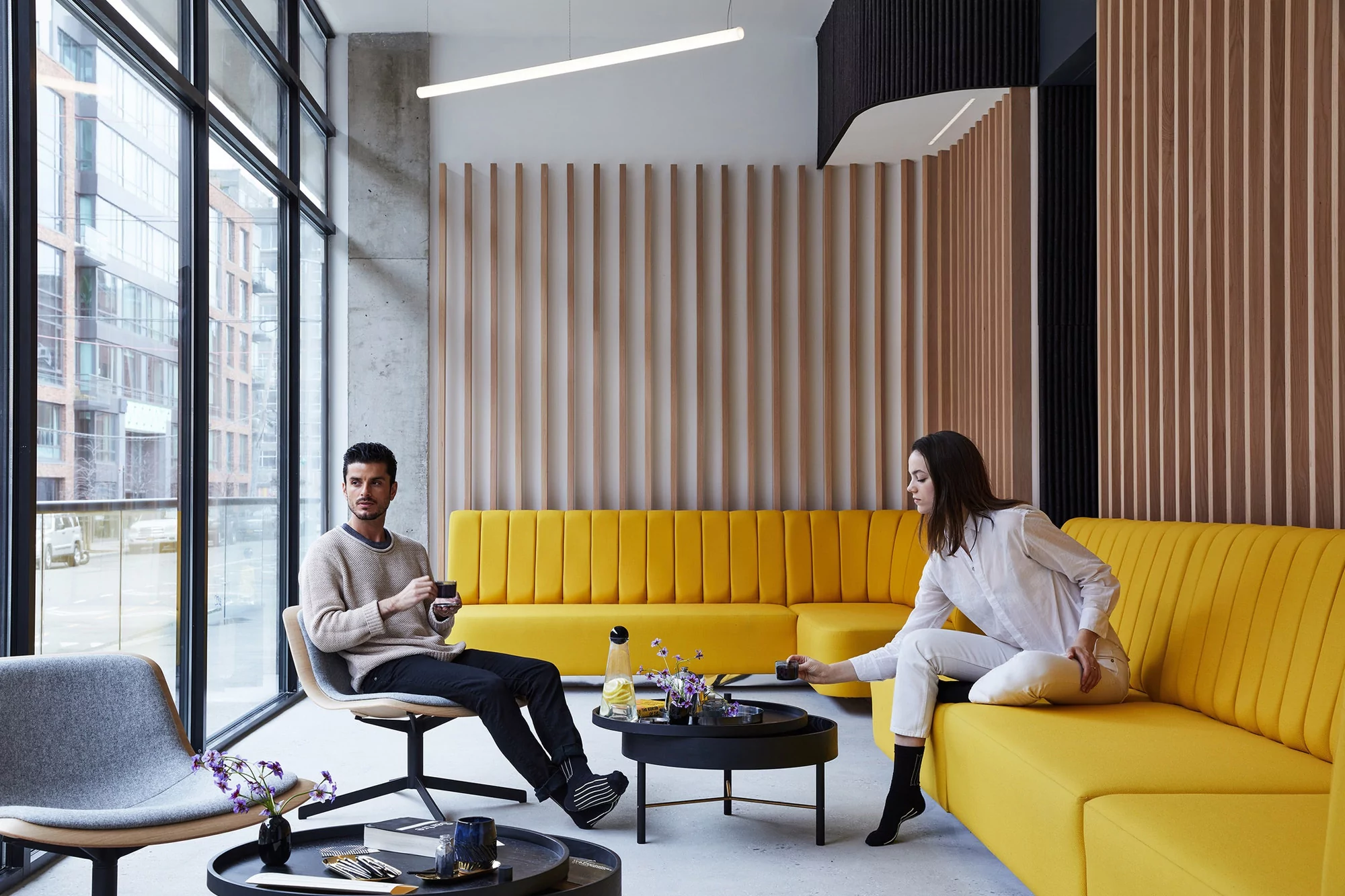 Two adults sitting in an indoor lounge area with yellow couches, modern furniture, wooden slat wall, and large windows showing city buildings.