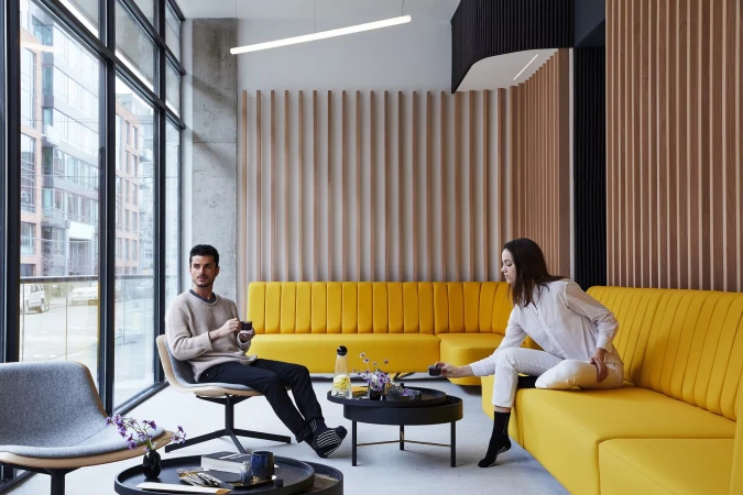 Two adults sitting in an indoor lounge area with yellow couches, modern furniture, wooden slat wall, and large windows showing city buildings.