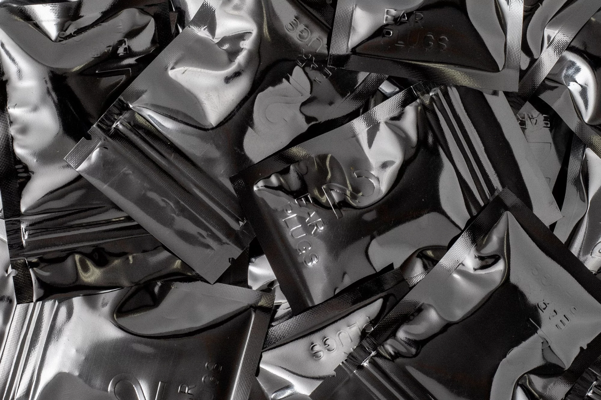 Pile of shiny black foil packets, stacked and slightly overlapping, filling the entire frame, with subtle embossed text on the surface.
