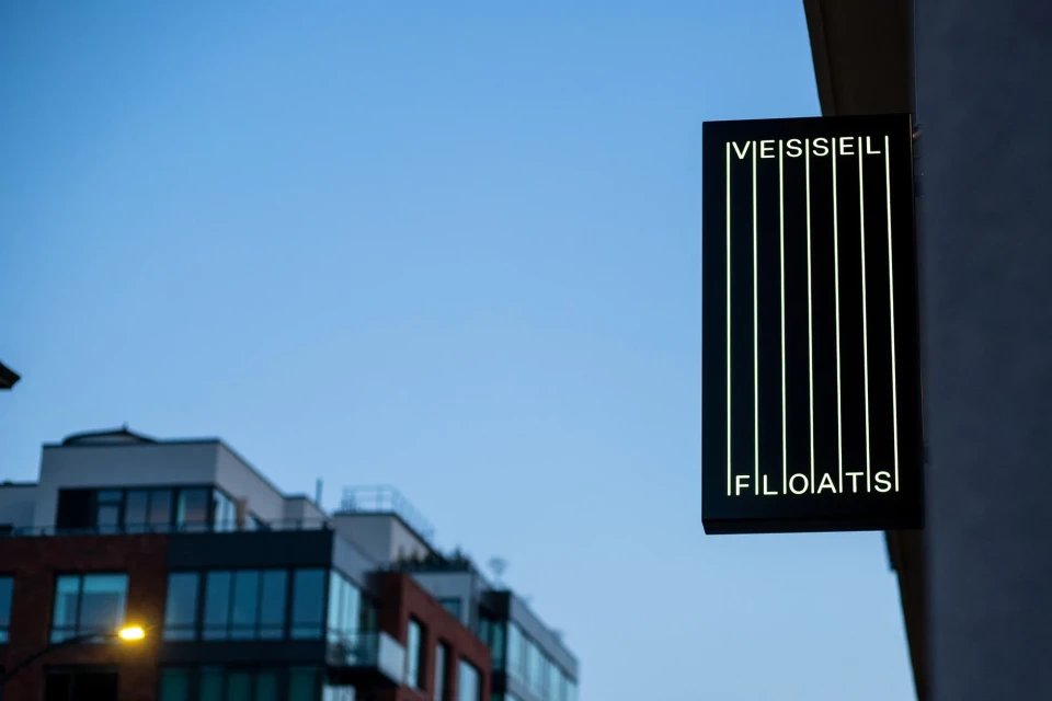 Building sign illuminated with the words 'Vessel Floats' against a dusk sky with apartment buildings in the soft-focus background.