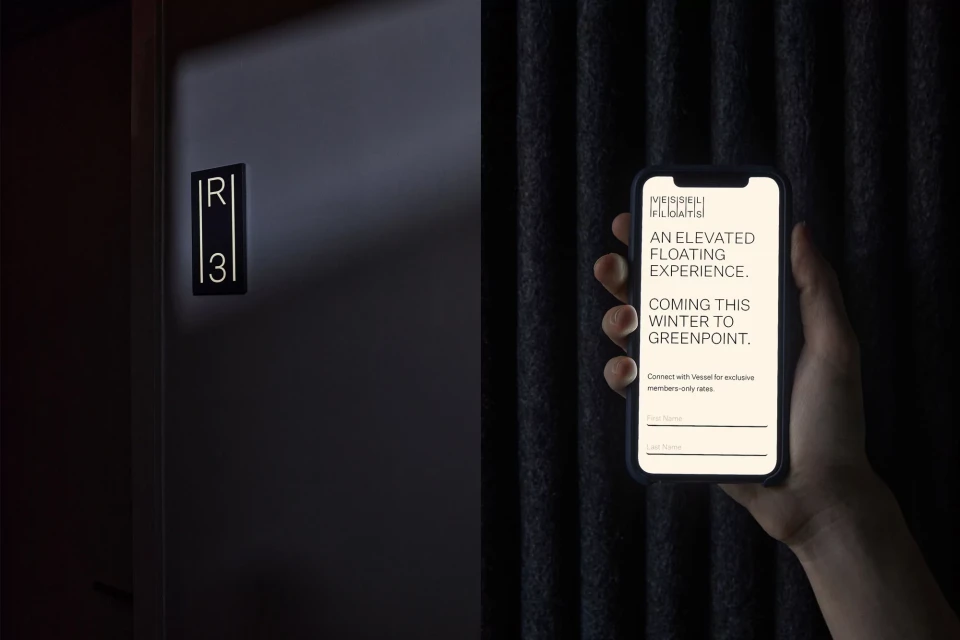 Hand holding smartphone showing text for Vessel Floats, advertising an elevated floating experience coming to Greenpoint this winter.