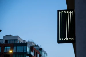 Illuminated rectangular hanging sign reading Vessel Floats against a twilight sky and modern building exterior