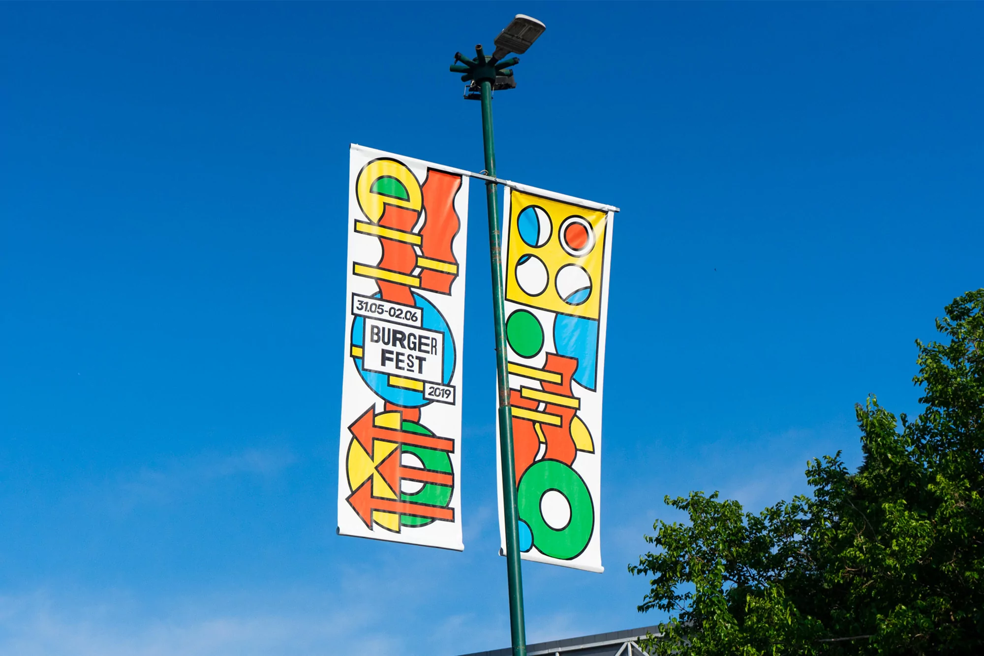 Two vertical banners hang from a lamppost against a blue sky, advertising the Burger Fest 2019 with colorful abstract graphics.