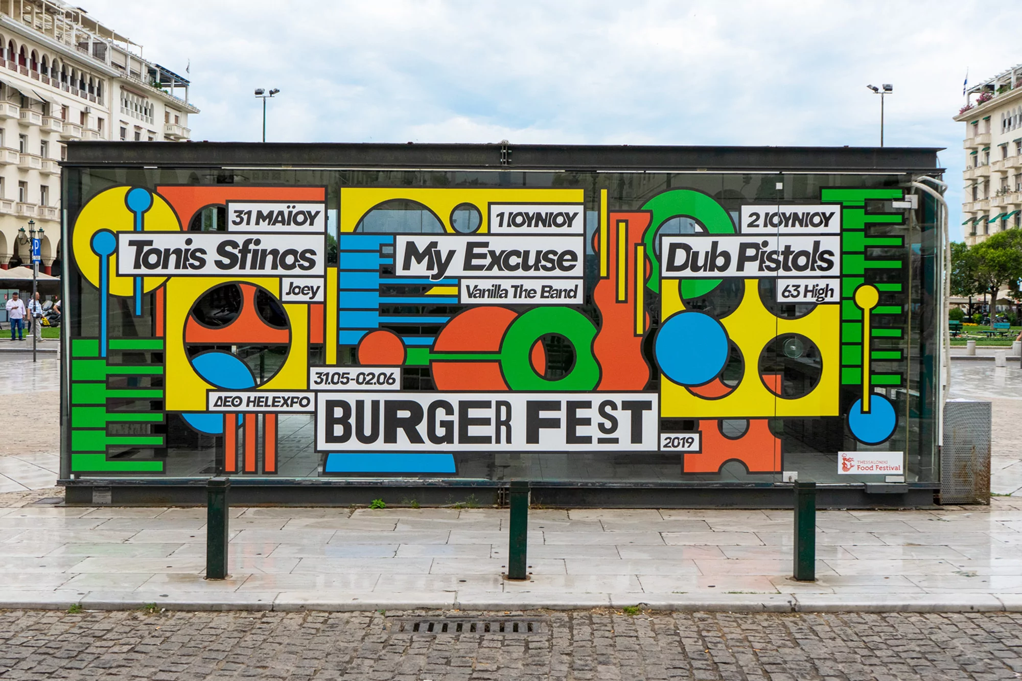 Outdoor view of a colorful Burger Fest advertisement in Thessaloniki, Greece, featuring band names, dates, and geometric shapes.