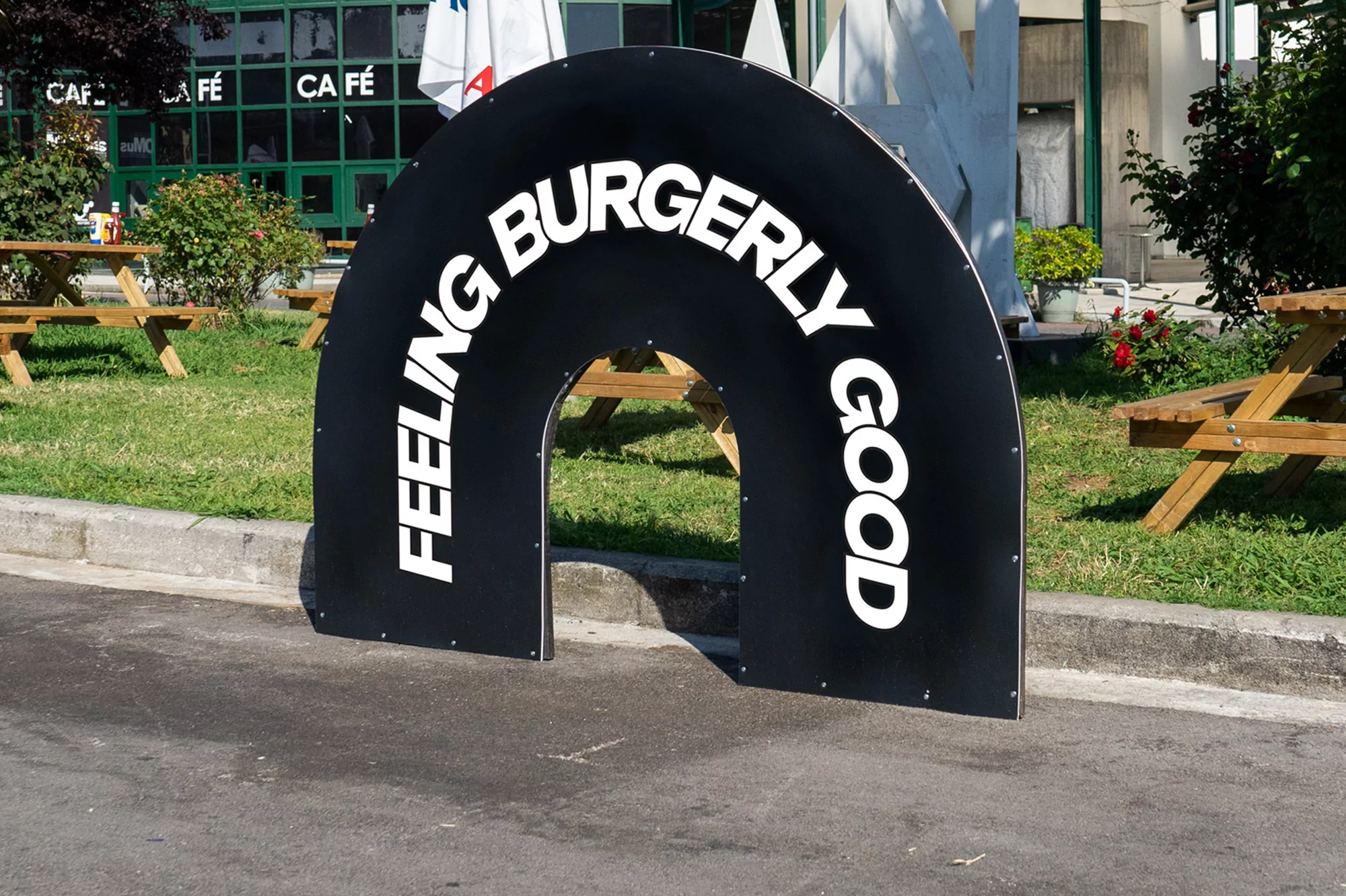 Black, arched, promotional sign with 'Feeling Burgerly Good' text, sits on pavement in front of a cafe and picnic tables.