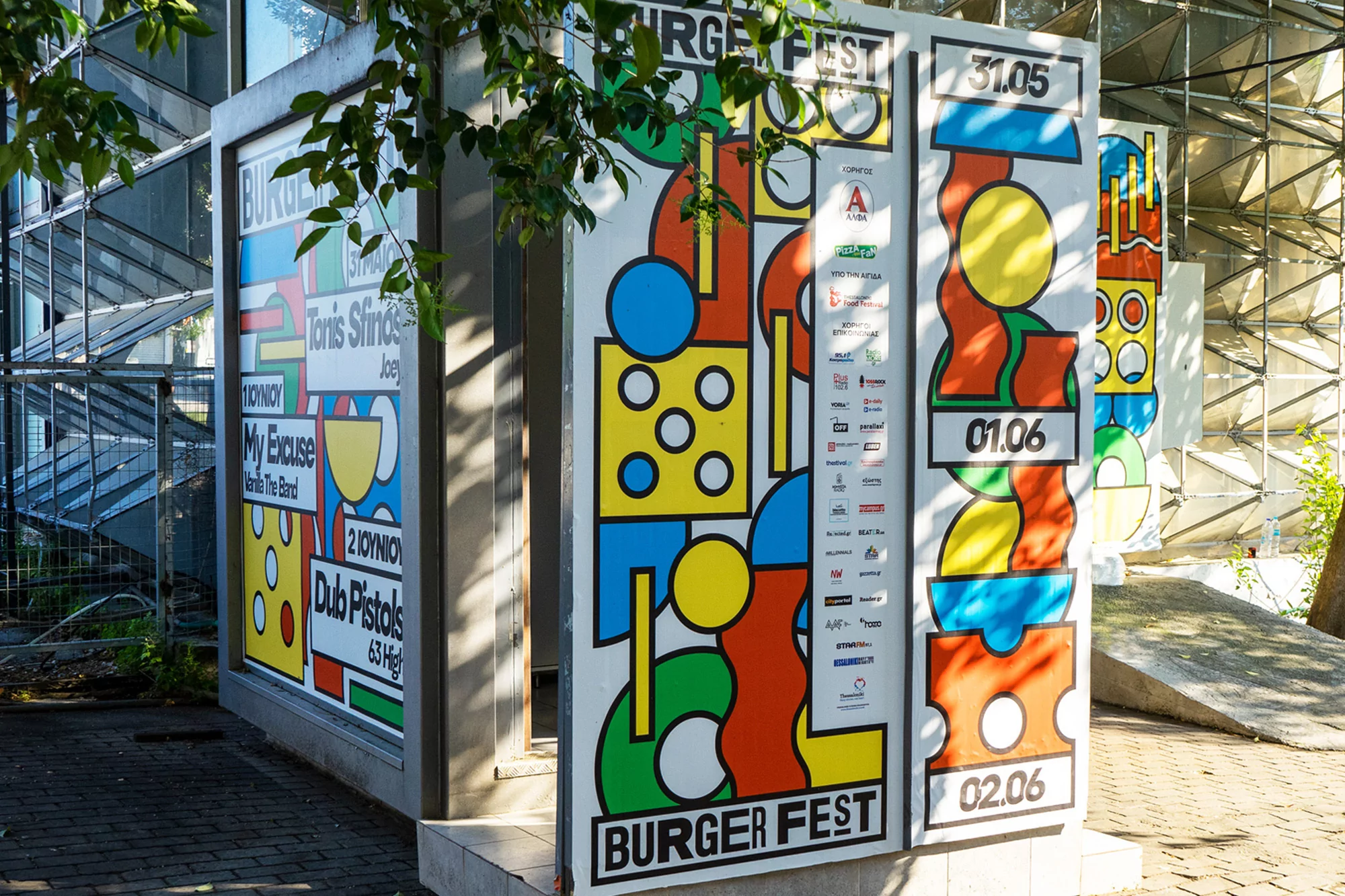 Colorful Burger Fest promotional posters with abstract graphics and event dates, stand outside an urban building under the shade of a tree.