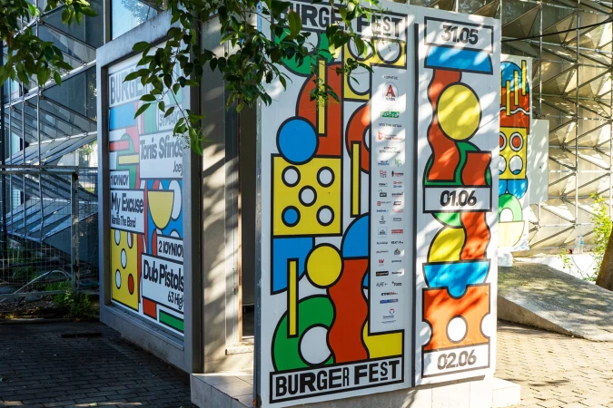Colorful Burger Fest promotional posters with abstract graphics and event dates, stand outside an urban building under the shade of a tree.