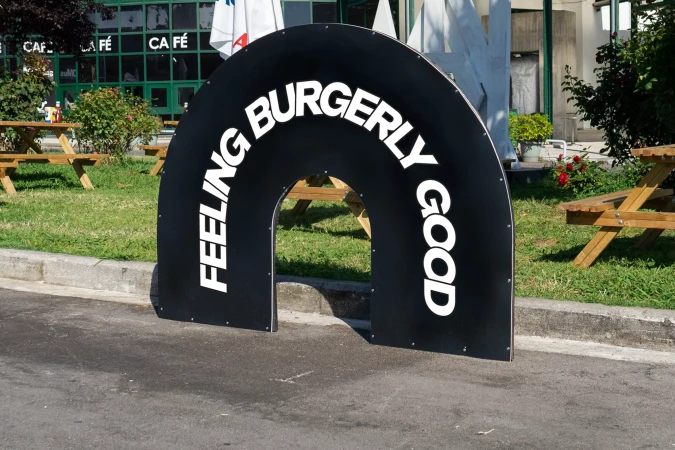 Black curved arch signage featuring white typography standing on asphalt next to a curb and a grassy lawn.
