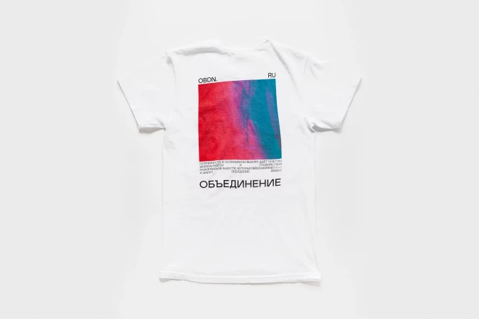 White t-shirt on white backdrop, square graphic in pink and teal with Russian text, simple studio photography for fashion brand.