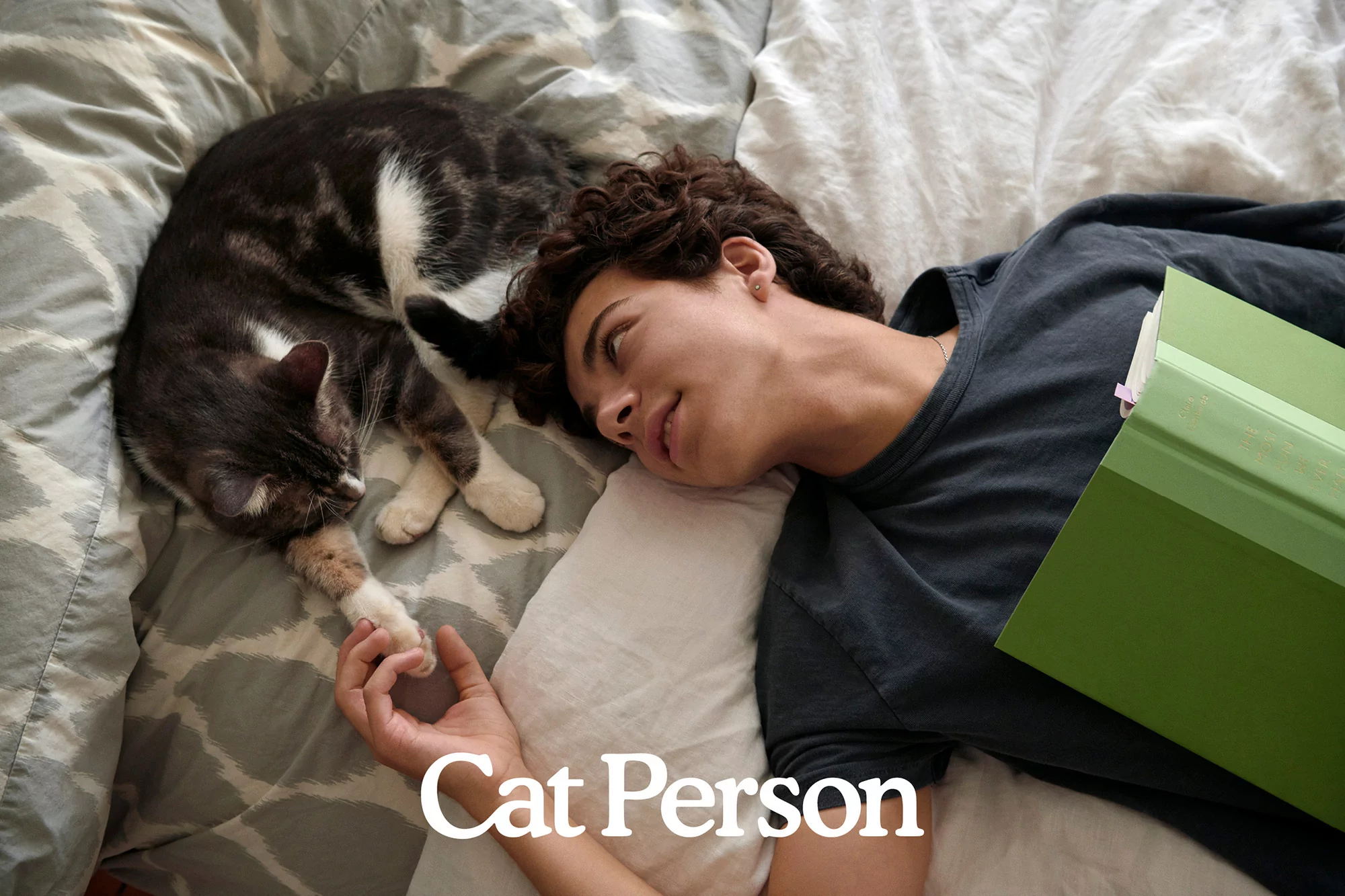 Overhead view of a young man lying on a bed with a grey tabby cat resting its paw on his hand; green book visible.