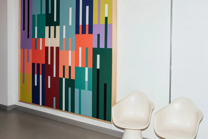 Abstract geometric art with rectangles of vibrant colors including red, green, and purple, hangs on a white wall with two white chairs.