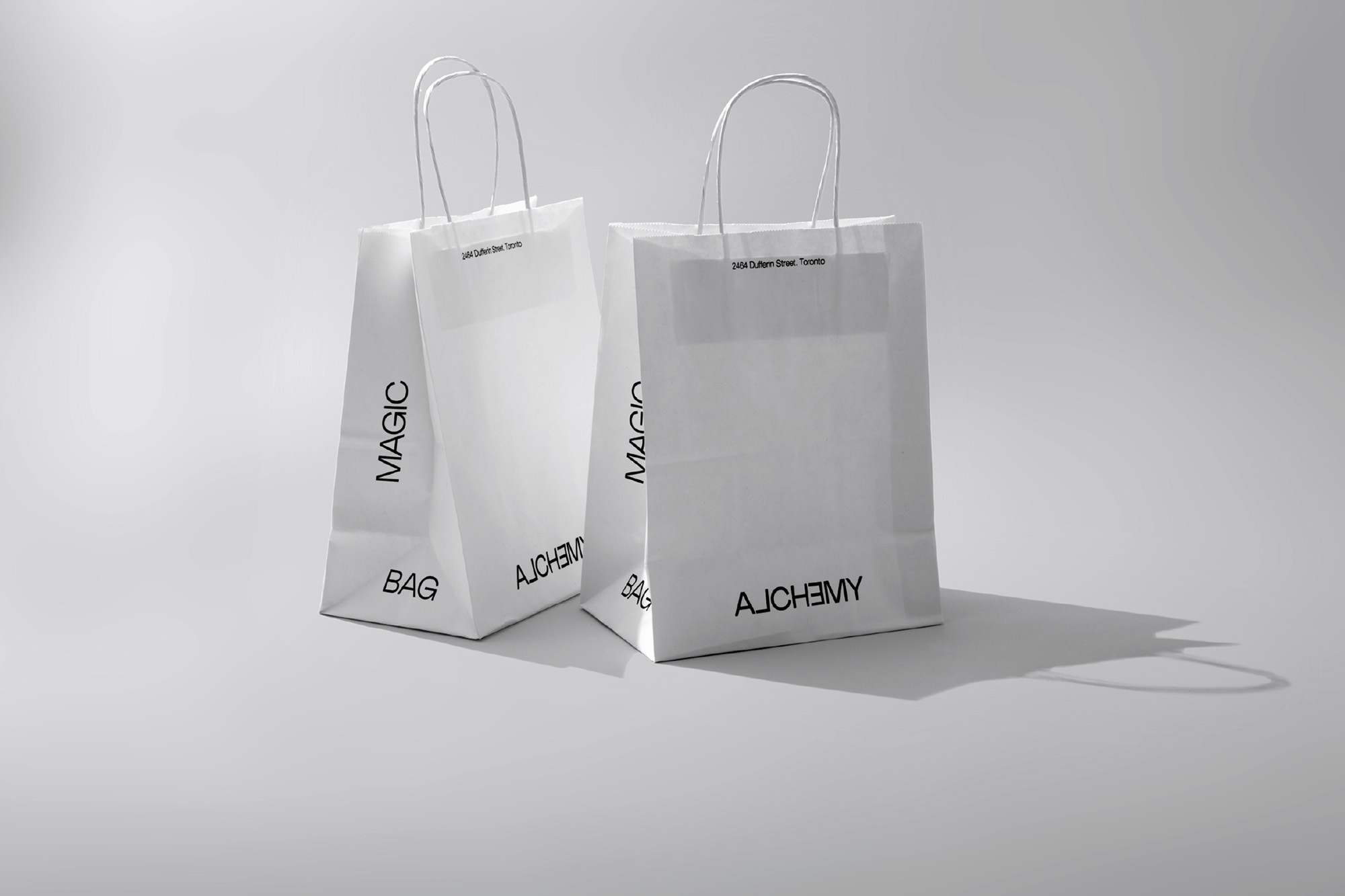 Two white paper shopping bags with rope handles and black text sit against a gradient white backdrop, casting soft shadows.