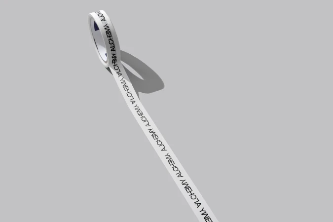 Close-up of a roll of white tape with the word 'ALCHEMY' printed in black in a repeating pattern on a gray background.