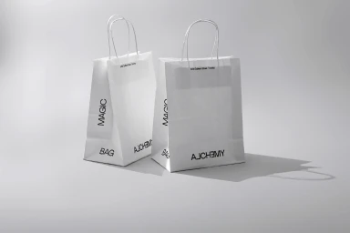 Two white paper shopping bags with black typography standing on a grey surface against a neutral background.