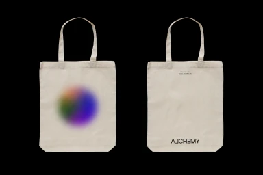 Front and back views of a cream canvas tote bag featuring a colorful blurred gradient orb and Alchemy branding against black.