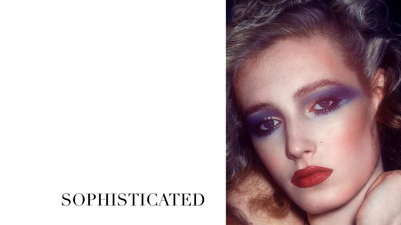 Close-up of a model showcasing dramatic purple and red makeup, highlighting her eyes and lips, against a white and dark backdrop.