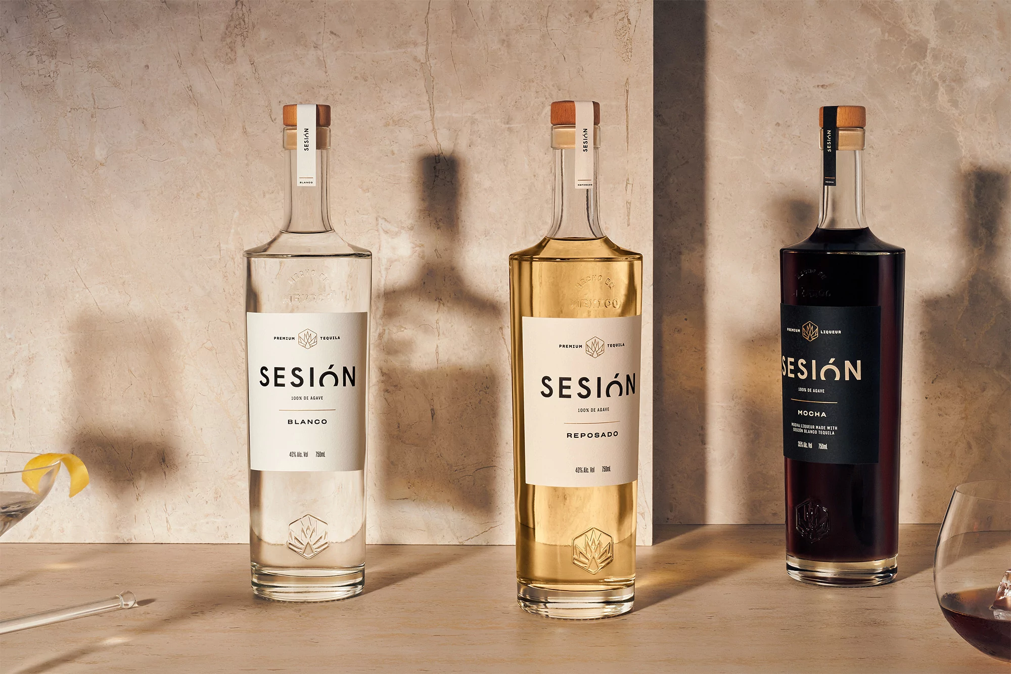 Three bottles of Sesión tequila, showcasing Blanco, Reposado, and Mocha varieties, on a neutral surface with cocktail and marble backdrop.
