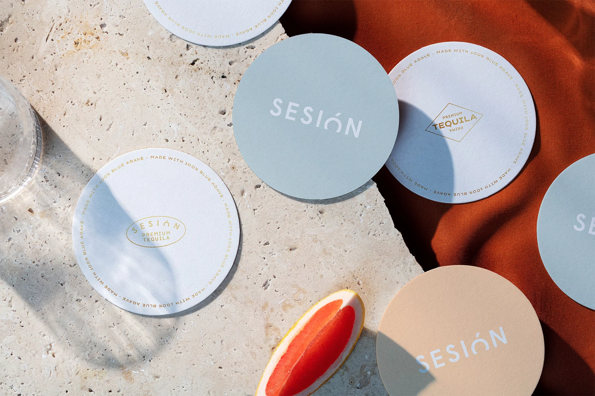 Studio shot of colorful 'Sesión' tequila coasters on a textured surface, featuring a grapefruit slice and soft, diffused lighting.