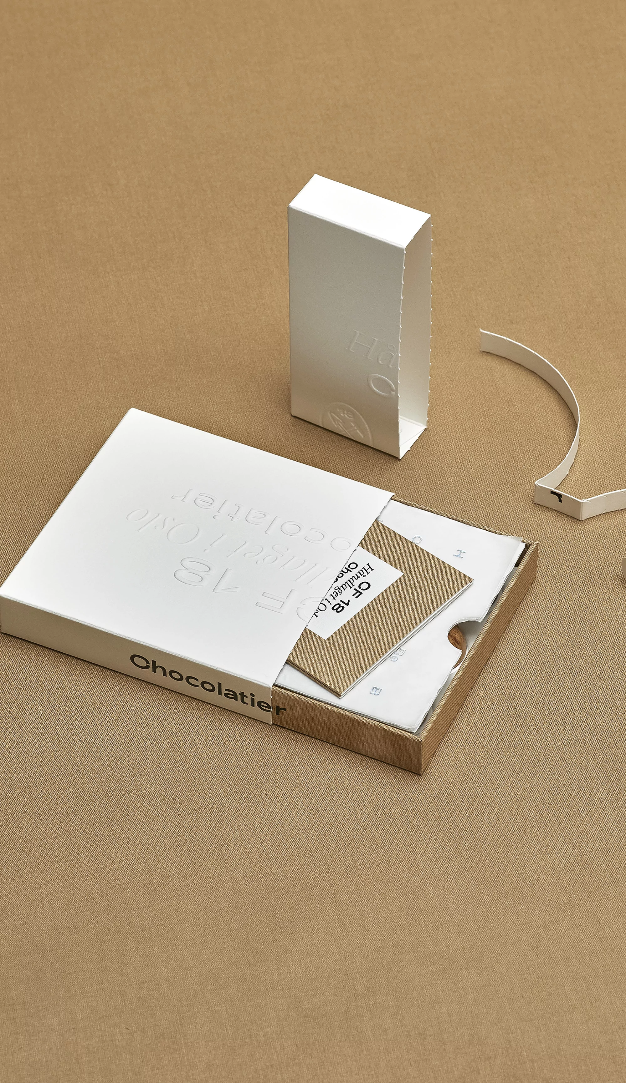 Angled studio shot of an open white chocolate box with a small white box on a tan background, presenting a modern minimalist aesthetic.