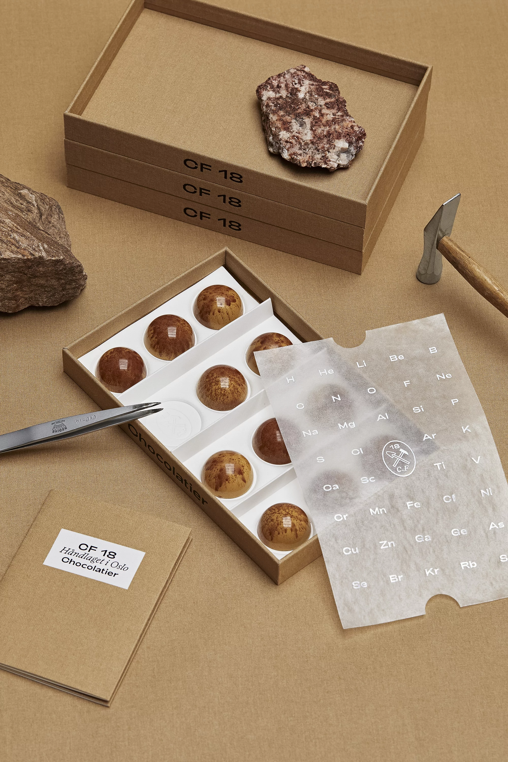 Artisanal chocolate bonbons in a box with a periodic table wrapper, presented with geological stone samples and a hammer on a neutral surface.
