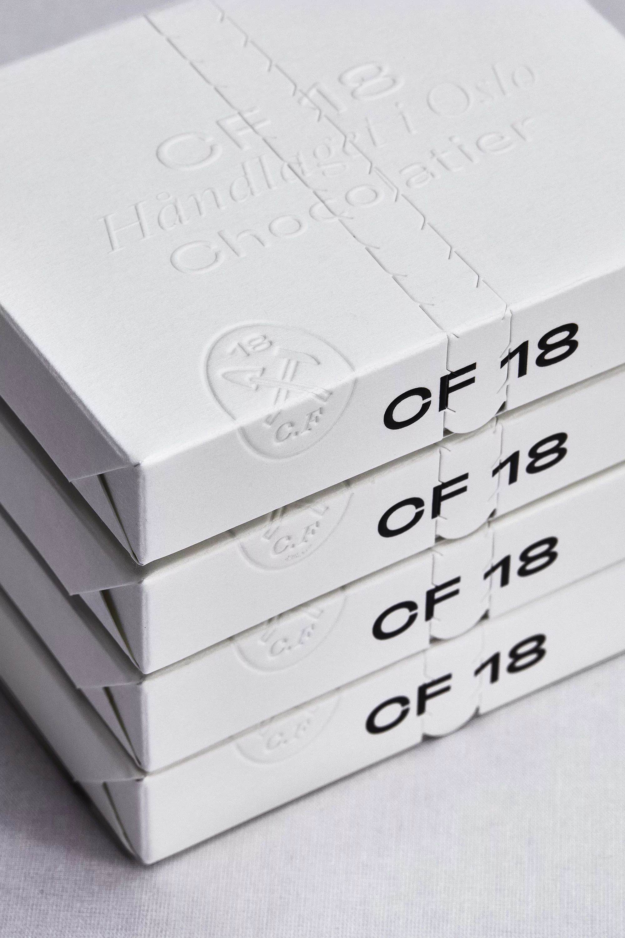 Four stacked white boxes with 'CF 18 Handlaget Oslo Chocolatier' text, wrapped in a ribbon, suggesting artisanal chocolate packaging.