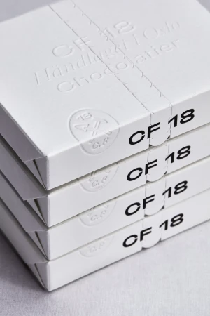 Four stacked white boxes with 'CF 18 Handlaget Oslo Chocolatier' text, wrapped in a ribbon, suggesting artisanal chocolate packaging.