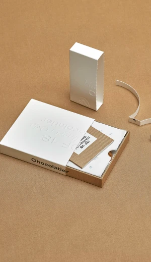 White paper packaging boxes with debossed typography on a brown textured paper surface by CF18 Chocolatier.
