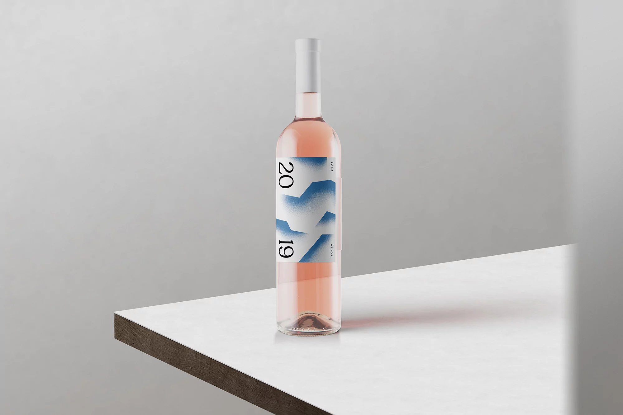 Bottle of rosé wine on a white table against a plain wall. Minimalist design with geometric shapes and '2019' printed on the label.