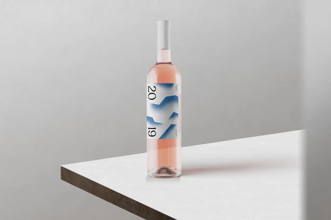 Bottle of rosé wine on a white table against a plain wall. Minimalist design with geometric shapes and '2019' printed on the label.