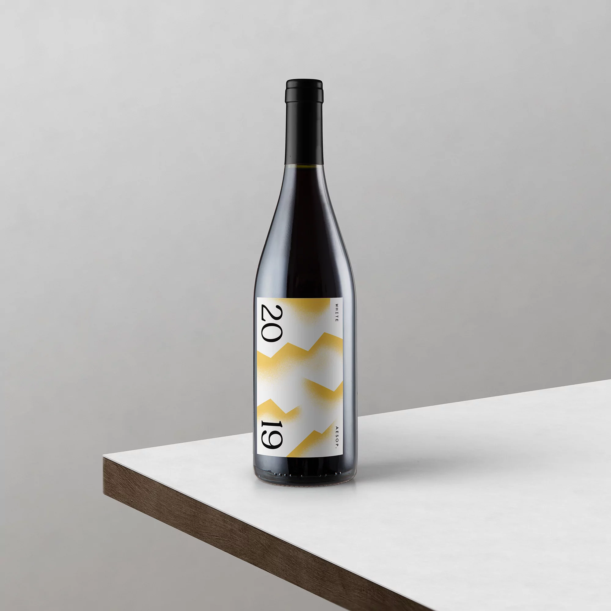 Dark red wine bottle with an abstract white and yellow label sits on a white surface against a plain gray background.