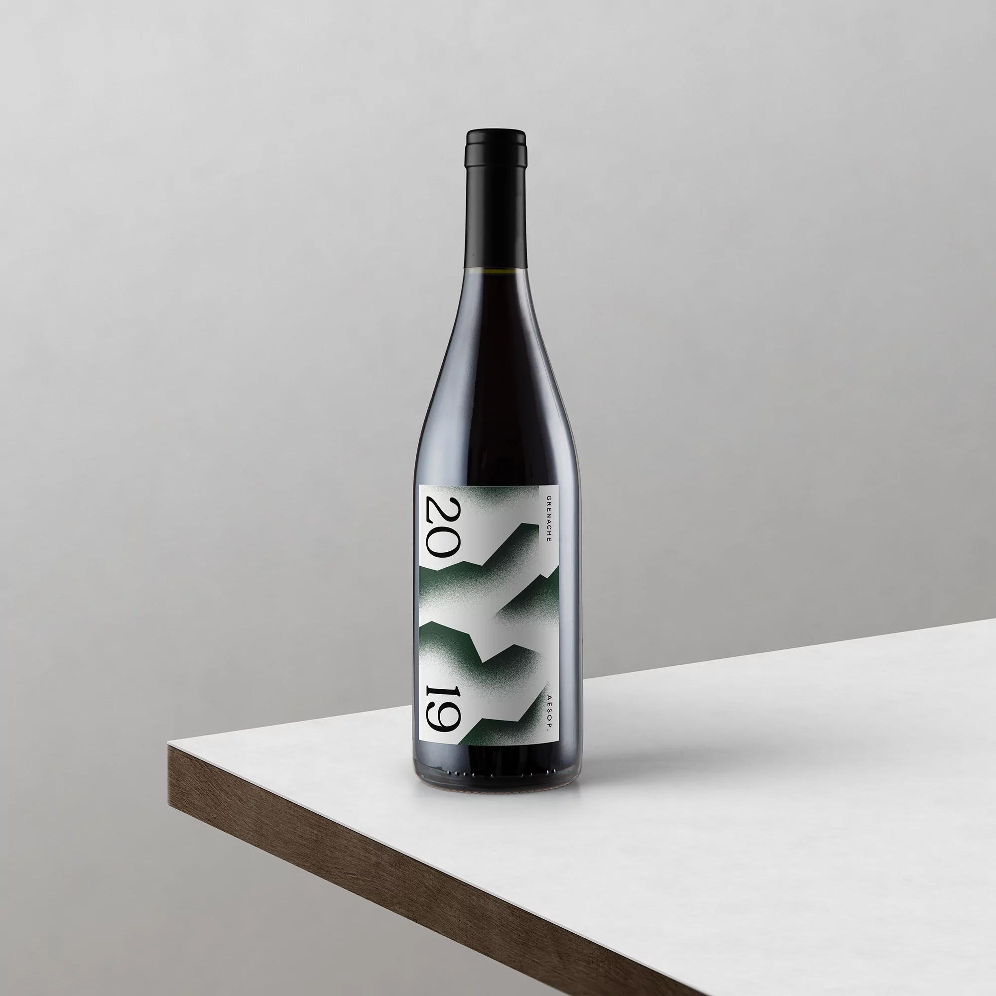 Studio shot of a bottle of red wine with a geometric label featuring a mountain-like design, against a white and grey background.