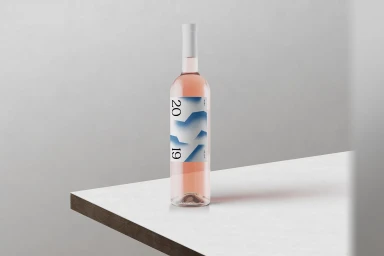 A bottle of rosé wine with a modern blue and white label standing on a white corner surface against a neutral background.