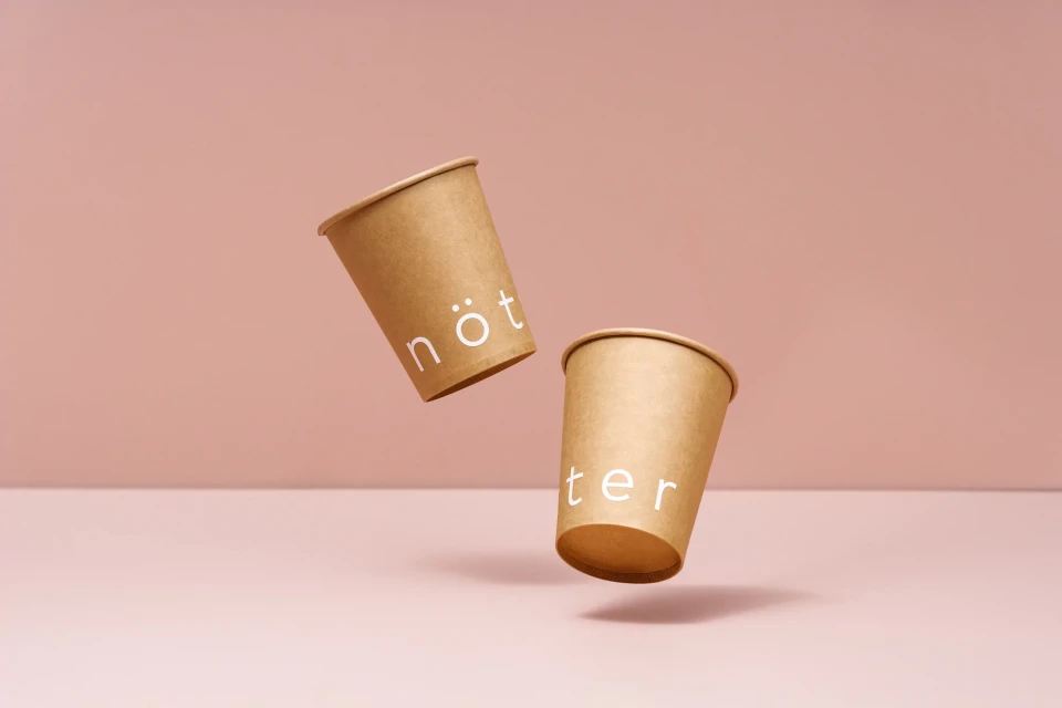 Two floating paper coffee cups with text stand against a pale pink backdrop, creating a minimalist and clean design.