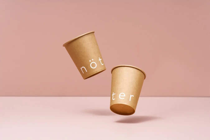 Two floating paper coffee cups with text stand against a pale pink backdrop, creating a minimalist and clean design.