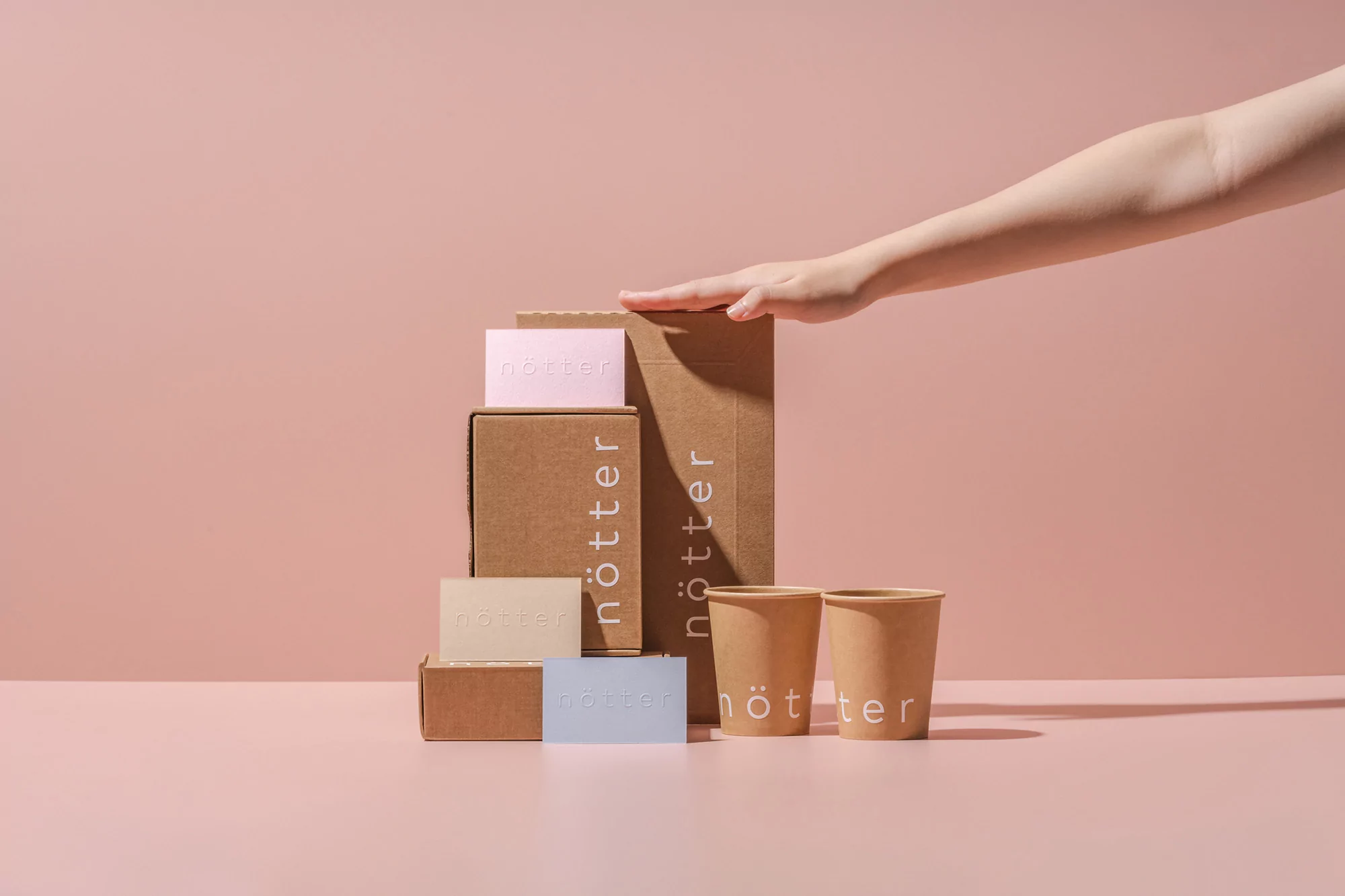 Hand reaches for a stacked set of cardboard boxes and paper cups, each printed with 'nötter' logo, on a pink background.