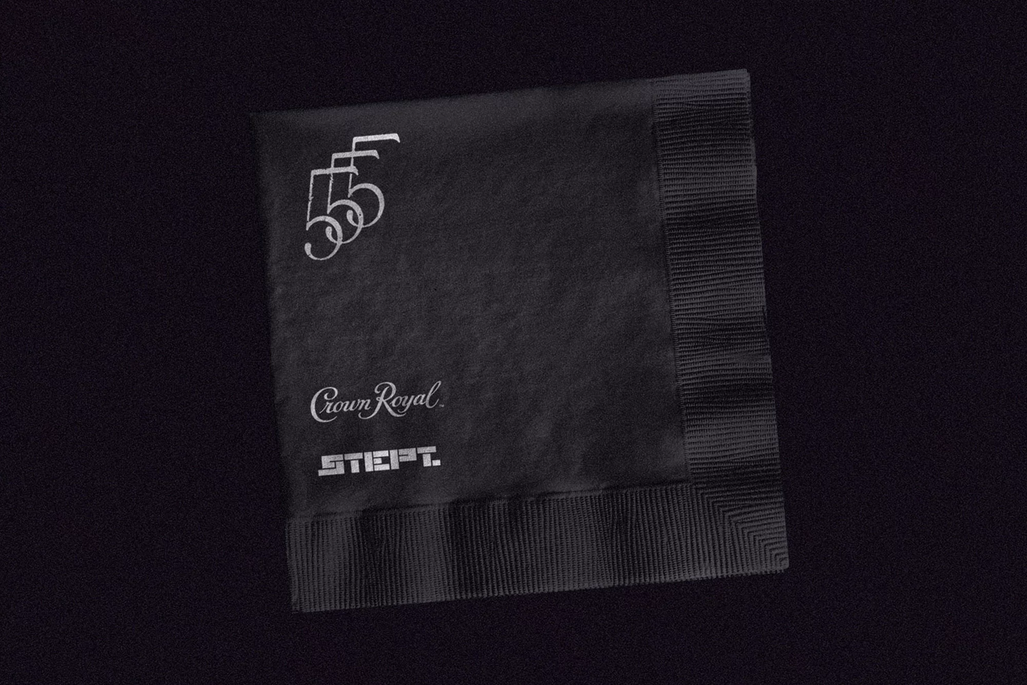 Black cocktail napkin with the logos and text of Crown Royal and STIEFT; placed on black fabric surface.