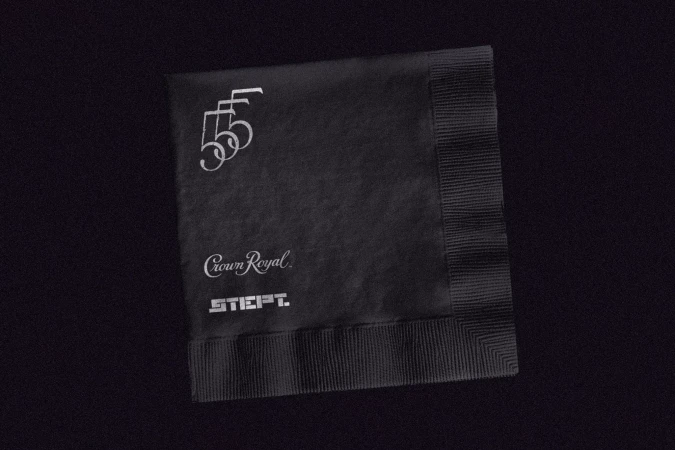 Black cocktail napkin with the logos and text of Crown Royal and STIEFT; placed on black fabric surface.