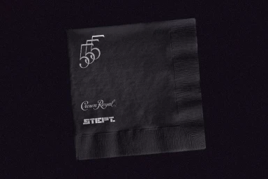 Black paper cocktail napkin featuring silver foil printed logos for Crown Royal and other branding against a dark background