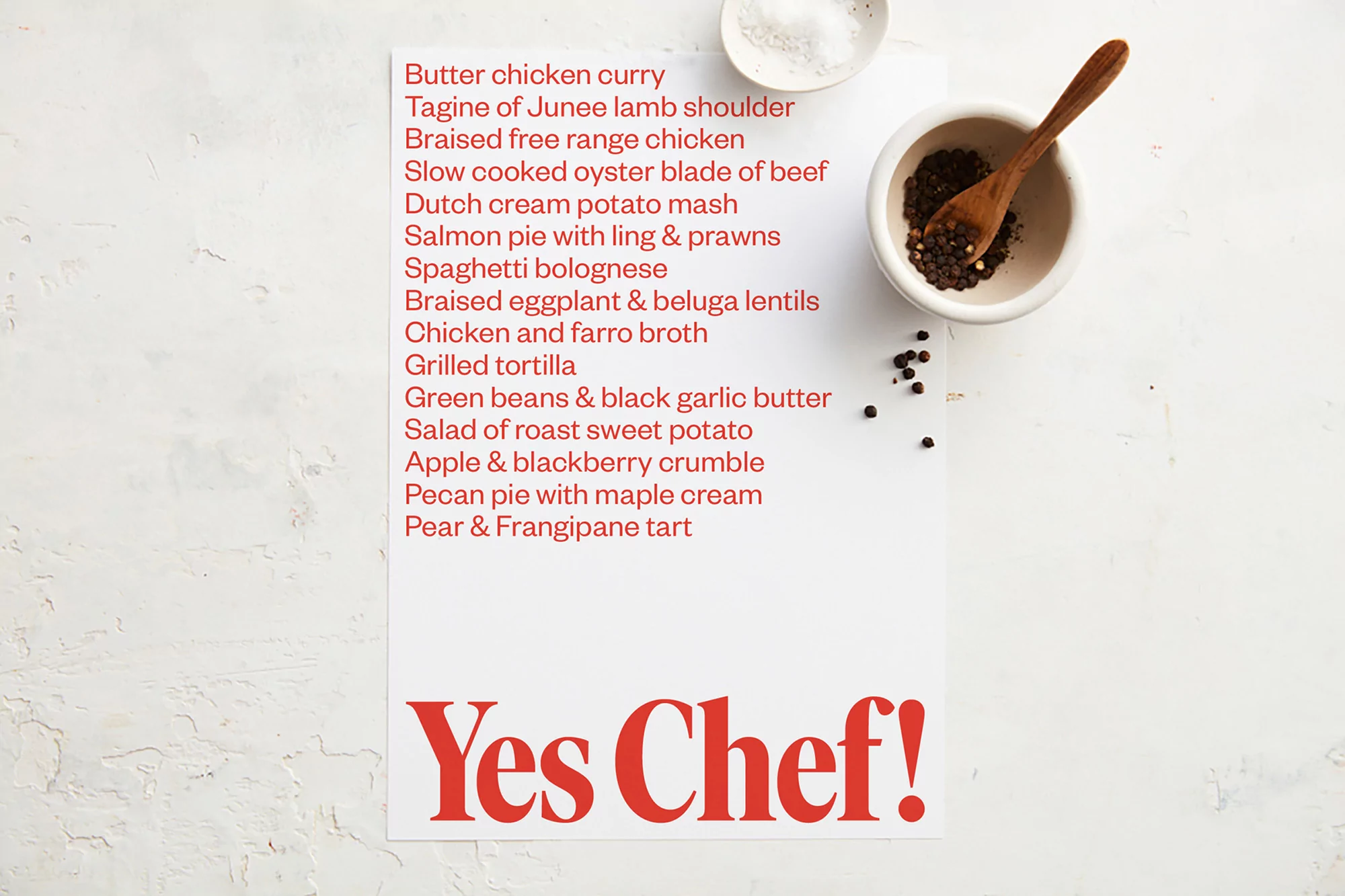 Overhead shot of a recipe list featuring various dishes alongside pepper grains and small bowls on a textured white surface.
