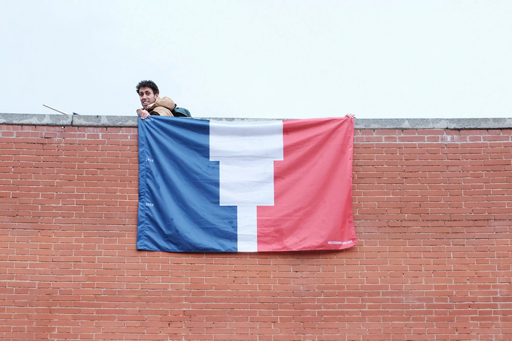 A person holding a flag with red, white, and blue stripes on a red brick wall outdoors on a cloudy day, smiling at the camera.