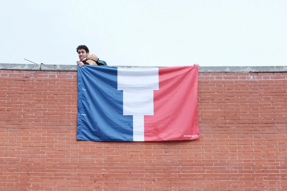 A person holding a flag with red, white, and blue stripes on a red brick wall outdoors on a cloudy day, smiling at the camera.