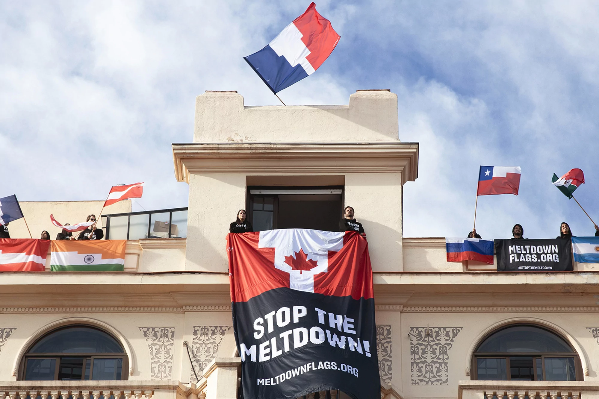 People hold flags of Canada, France, Chile, and other countries from a building with a banner that reads: Stop the Meltdown!