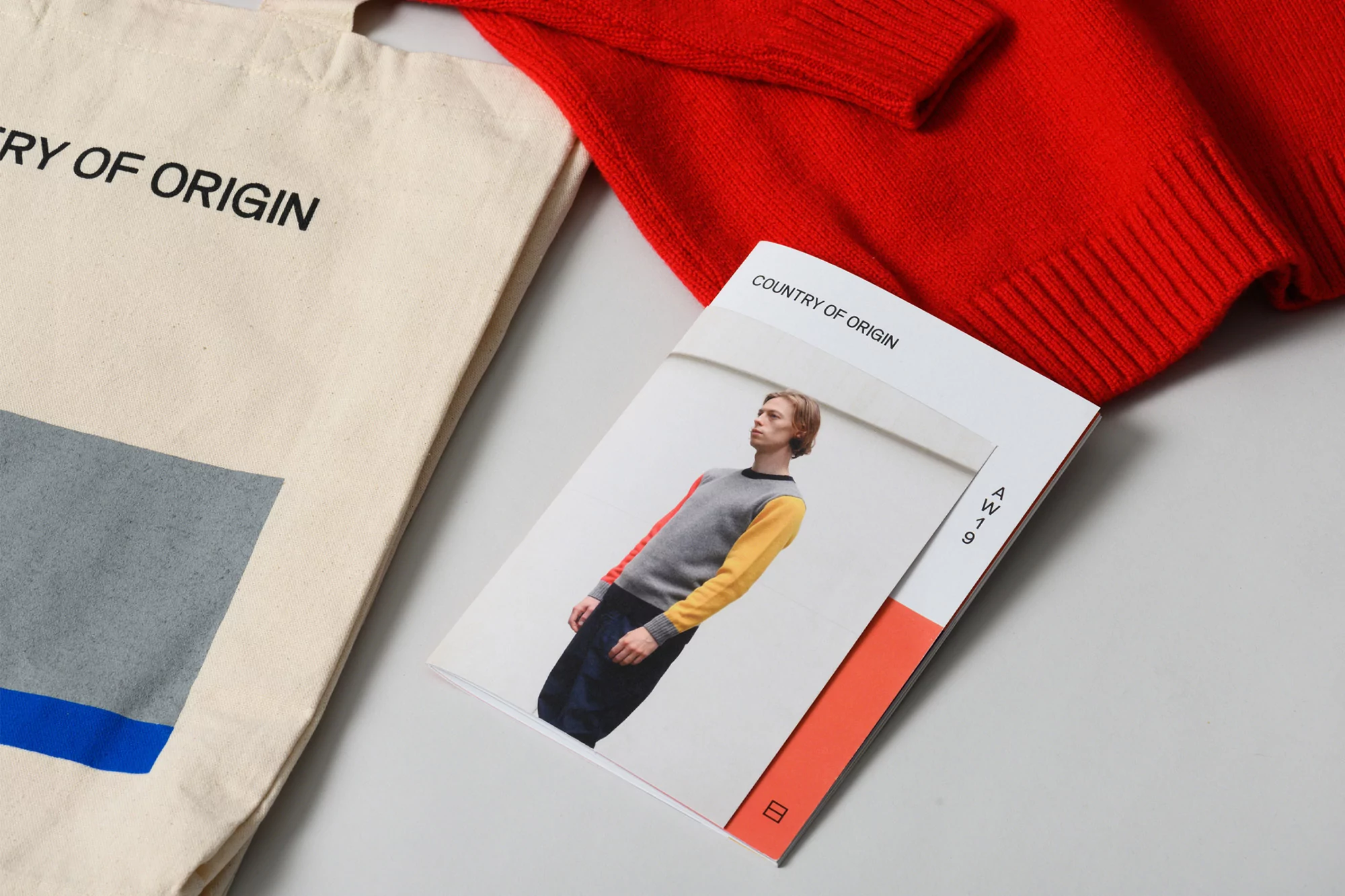 Still life of a red sweater, beige tote bag, and fashion catalog, showcasing Country of Origin brand on a plain background.