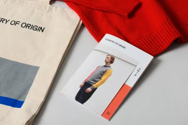 Top-down view of branded merchandise including a canvas tote bag, a red knit sweater, and a lookbook pamphlet on a neutral grey background.