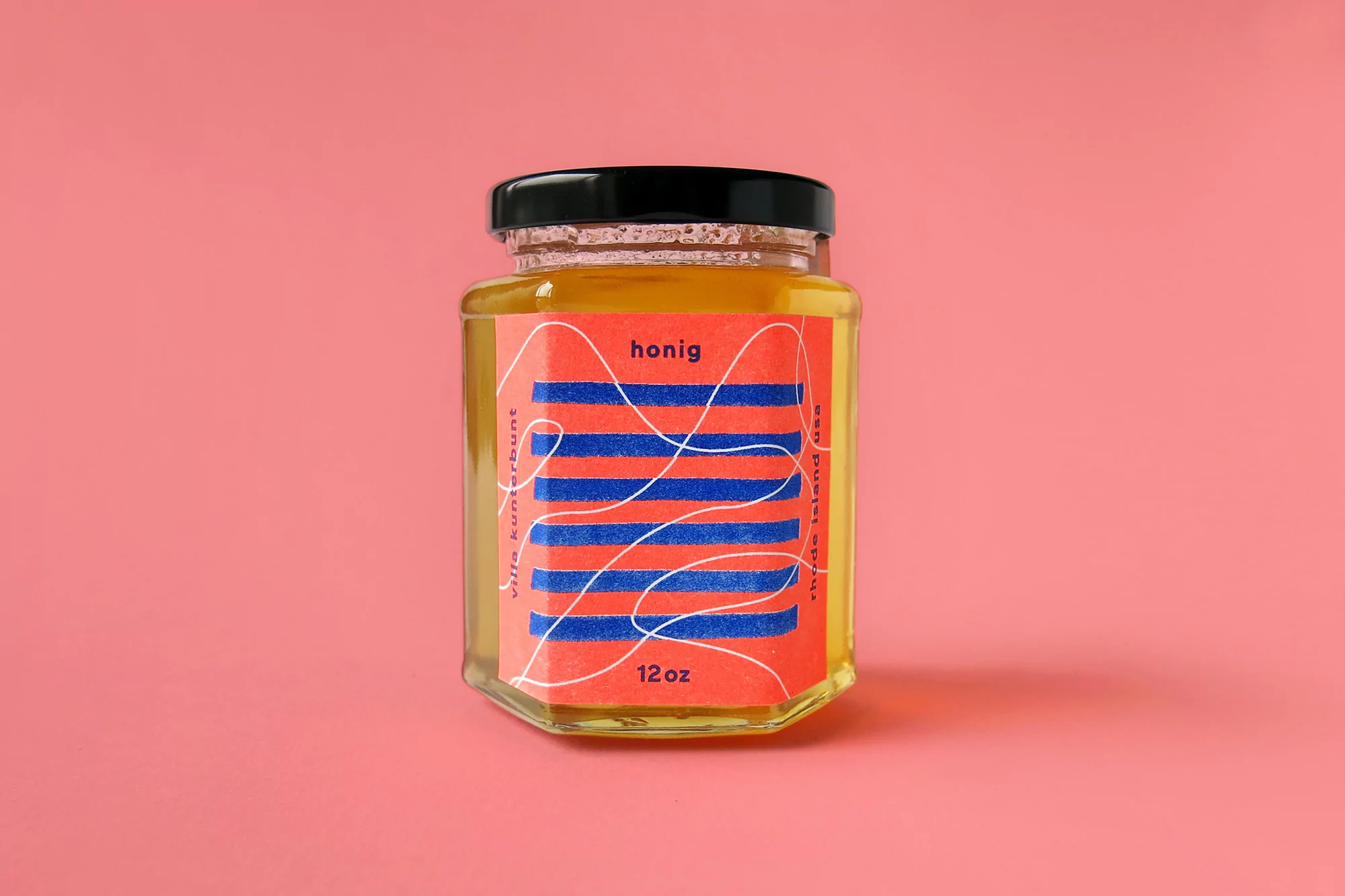 Glass jar of honey with a red and blue patterned label set against a plain pink background.