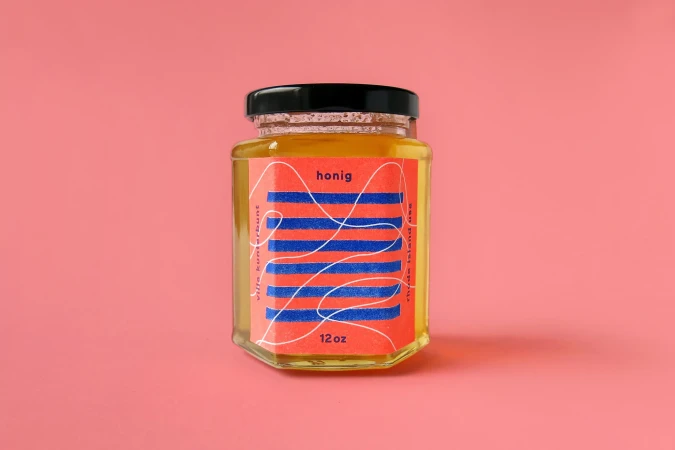 Glass jar of honey with a red and blue patterned label set against a plain pink background.